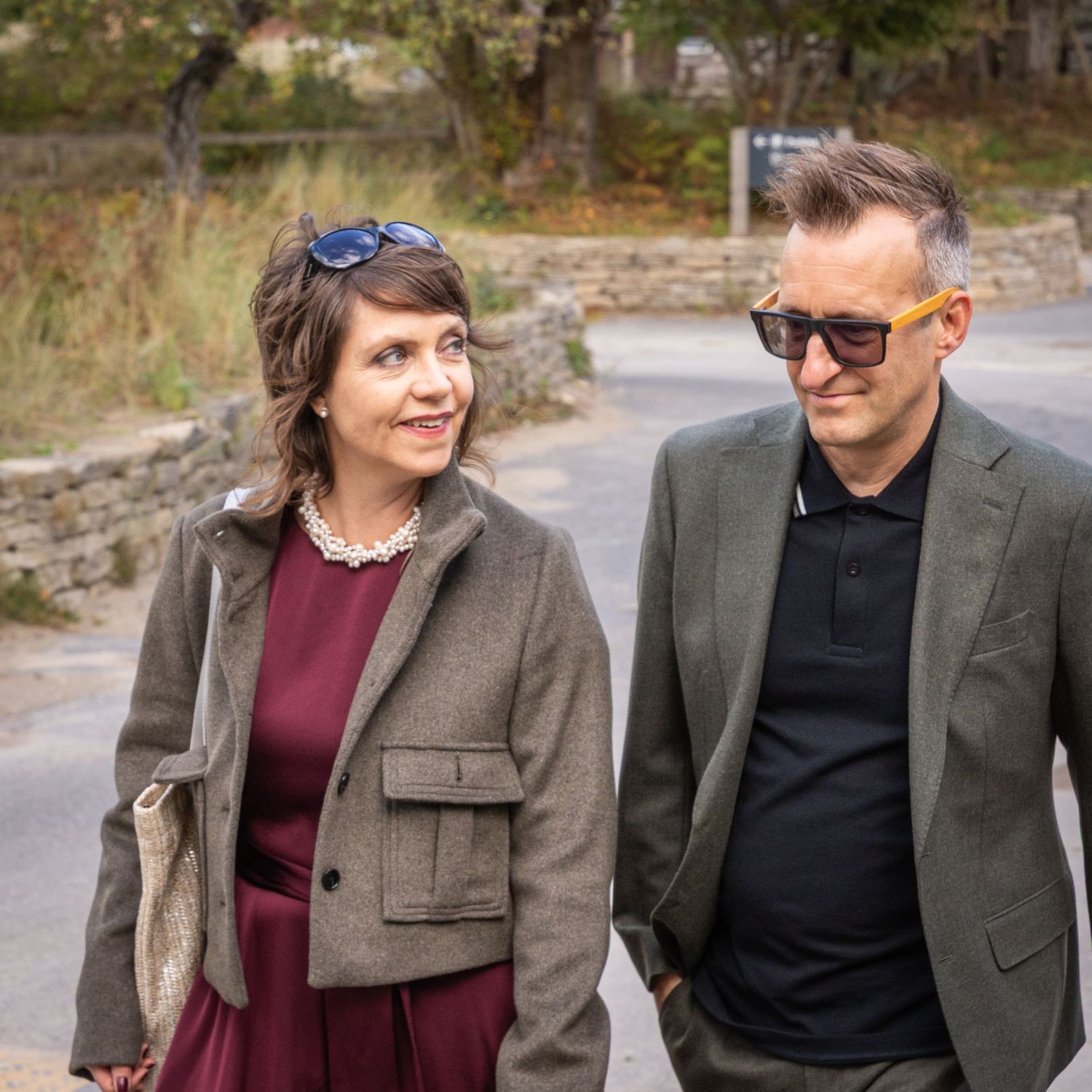 Couple in elegant attire walking outdoors, woman in burgundy dress and man in gray blazer with sunglasses.