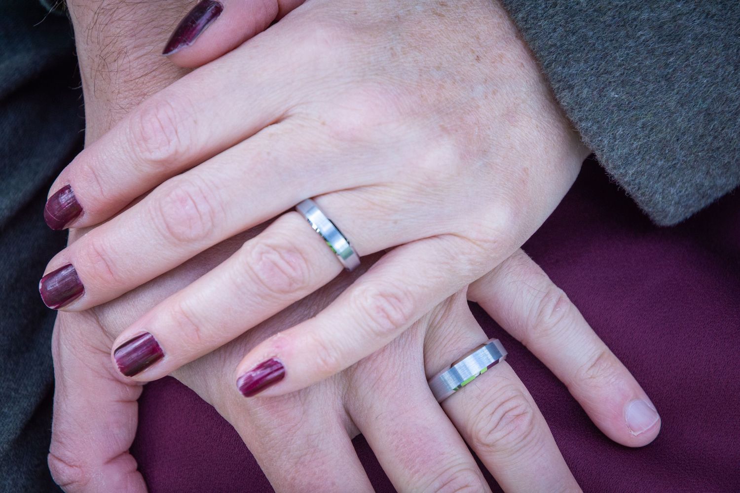Two hands wearing matching silver wedding bands, one with dark red nail polish, resting together symbolizing marriage.