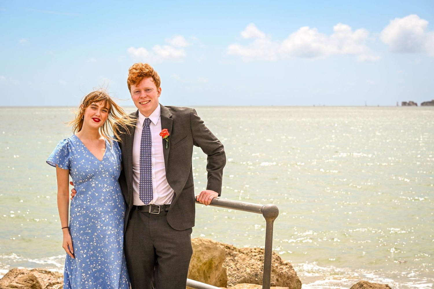 Couple posing by the seaside, woman in blue floral dress and man in dark suit with red flower, on a sunny day.