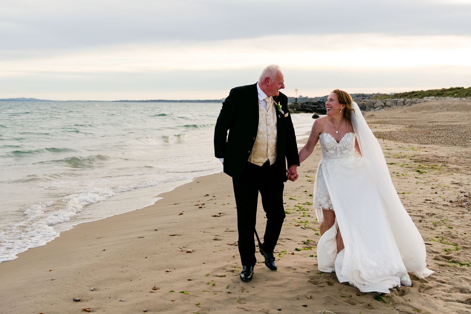 Bride and groom walking hand in hand on a sandy beach at sunset in wedding attire.