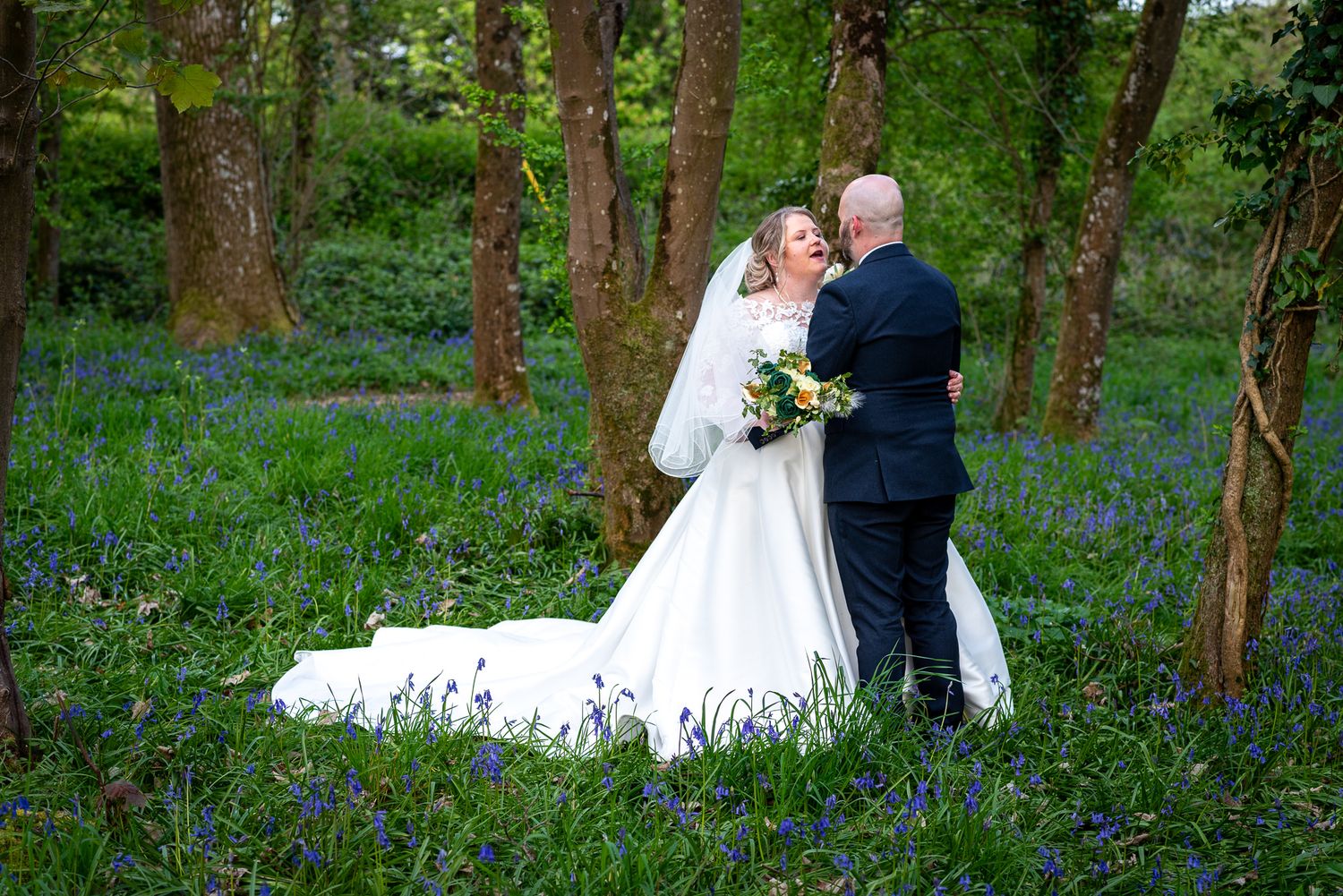 Bride and groom embracing in a bluebell woodland, bride in white gown holding bouquet, surrounded by lush green trees.
