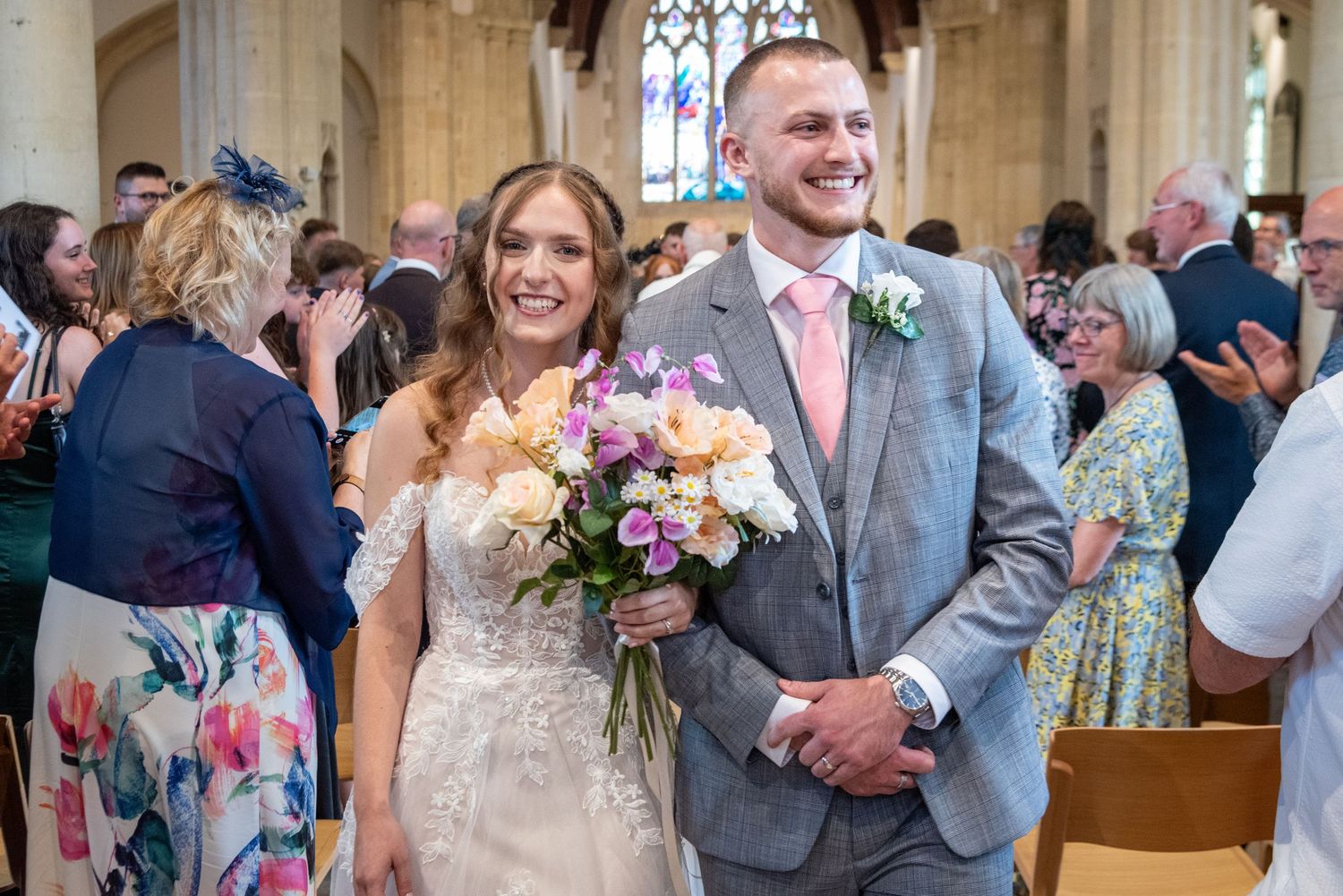 Bride and groom walking down the aisle smiling, surrounded by applauding wedding guests in a church.
