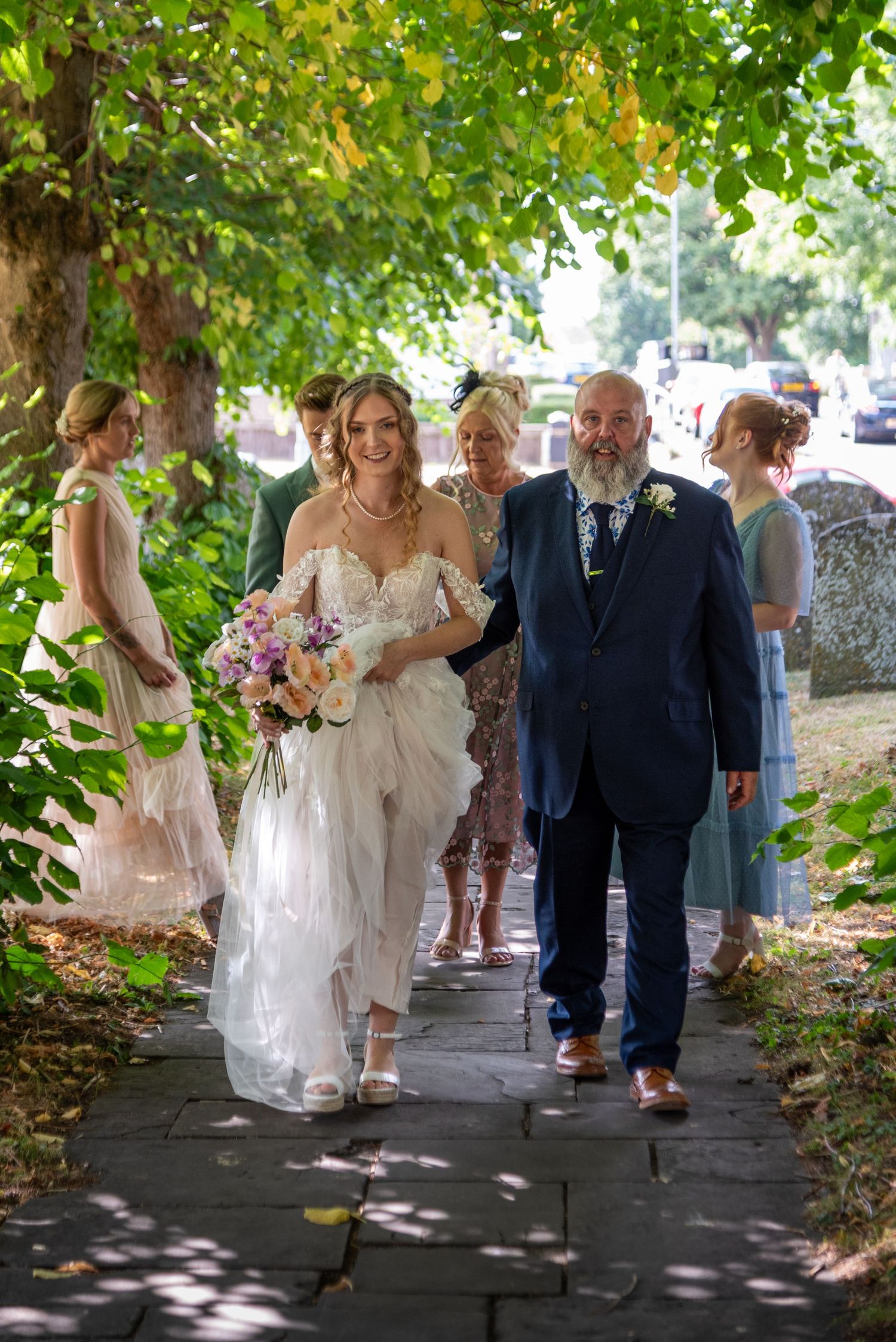 Bride walks down outdoor church path with bearded father in navy suit, bridesmaids following, holding colorful bouquet.