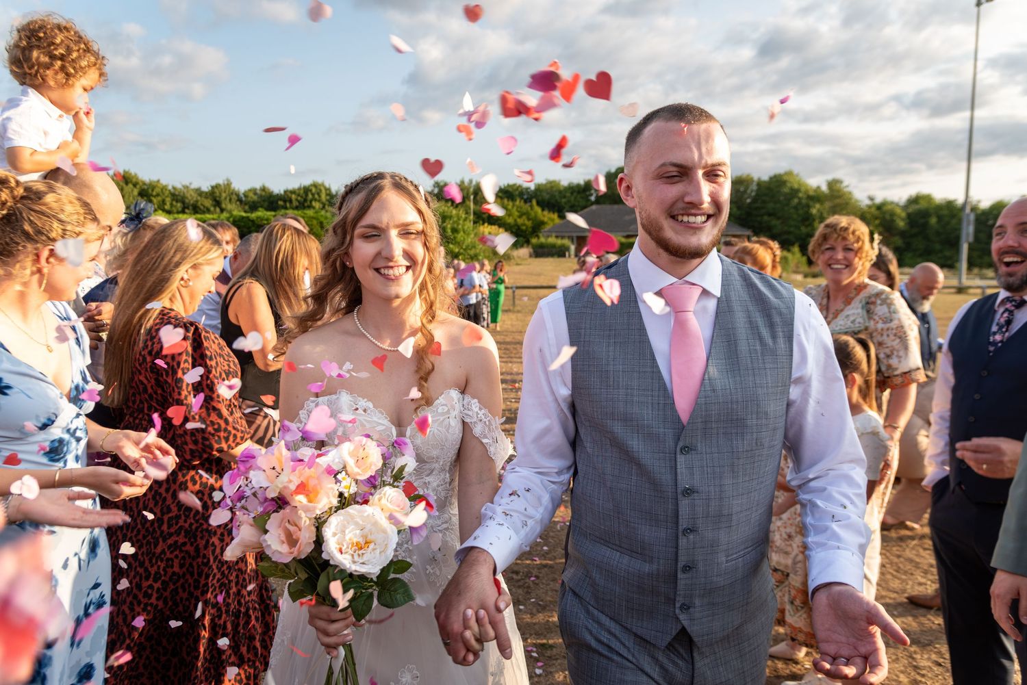 Bride and groom walk through confetti shower, smiling, as guests celebrate their wedding outdoors at golden hour.
