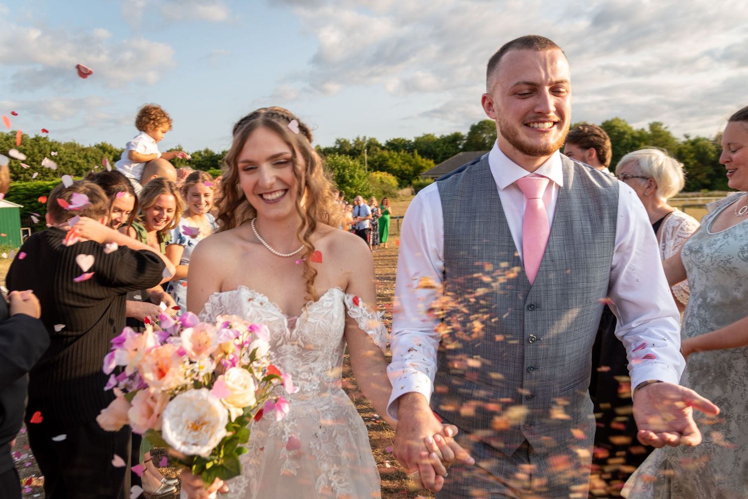 Wedding guests celebrate with confetti during an outdoor ceremony under a cloudy sky.