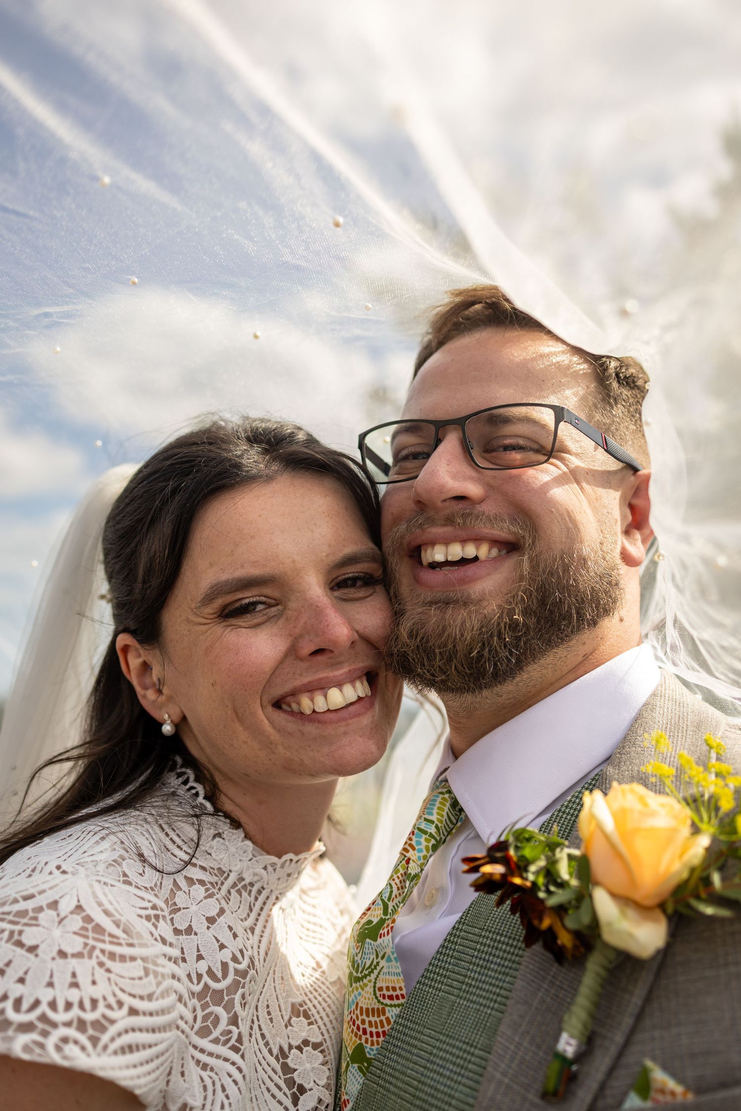 Happy newlyweds smiling under bridal veil against blue sky, groom wearing glasses and boutonniere.
