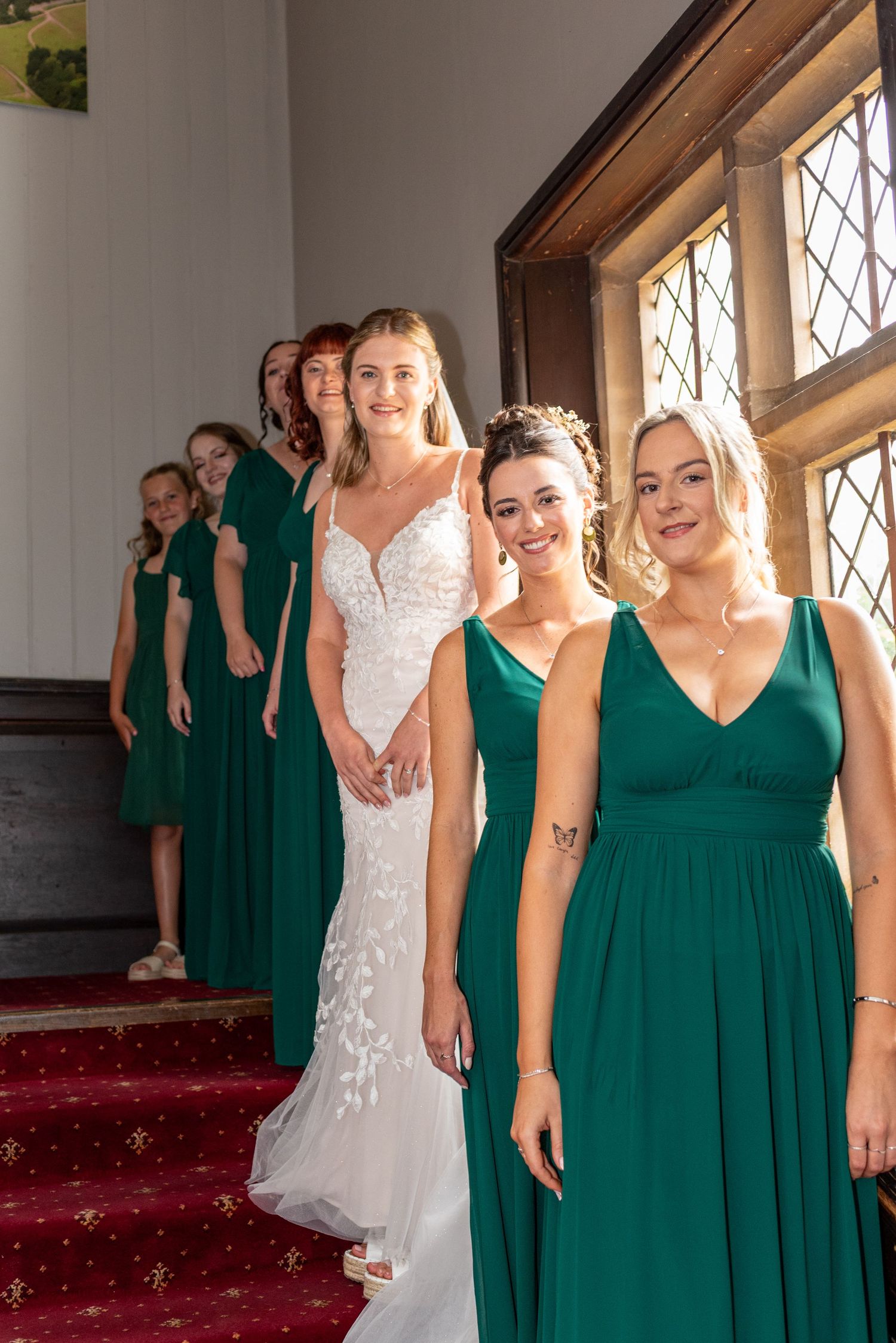 Bride in white gown stands with bridesmaids in emerald green dresses on red carpeted stairs near leaded windows.