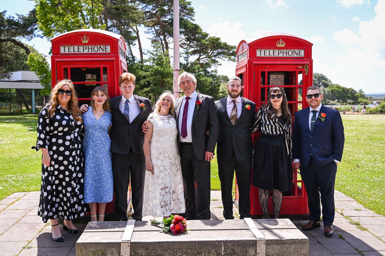 Group of eight people posing together outdoors near two iconic red British telephone boxes on a sunny day.
