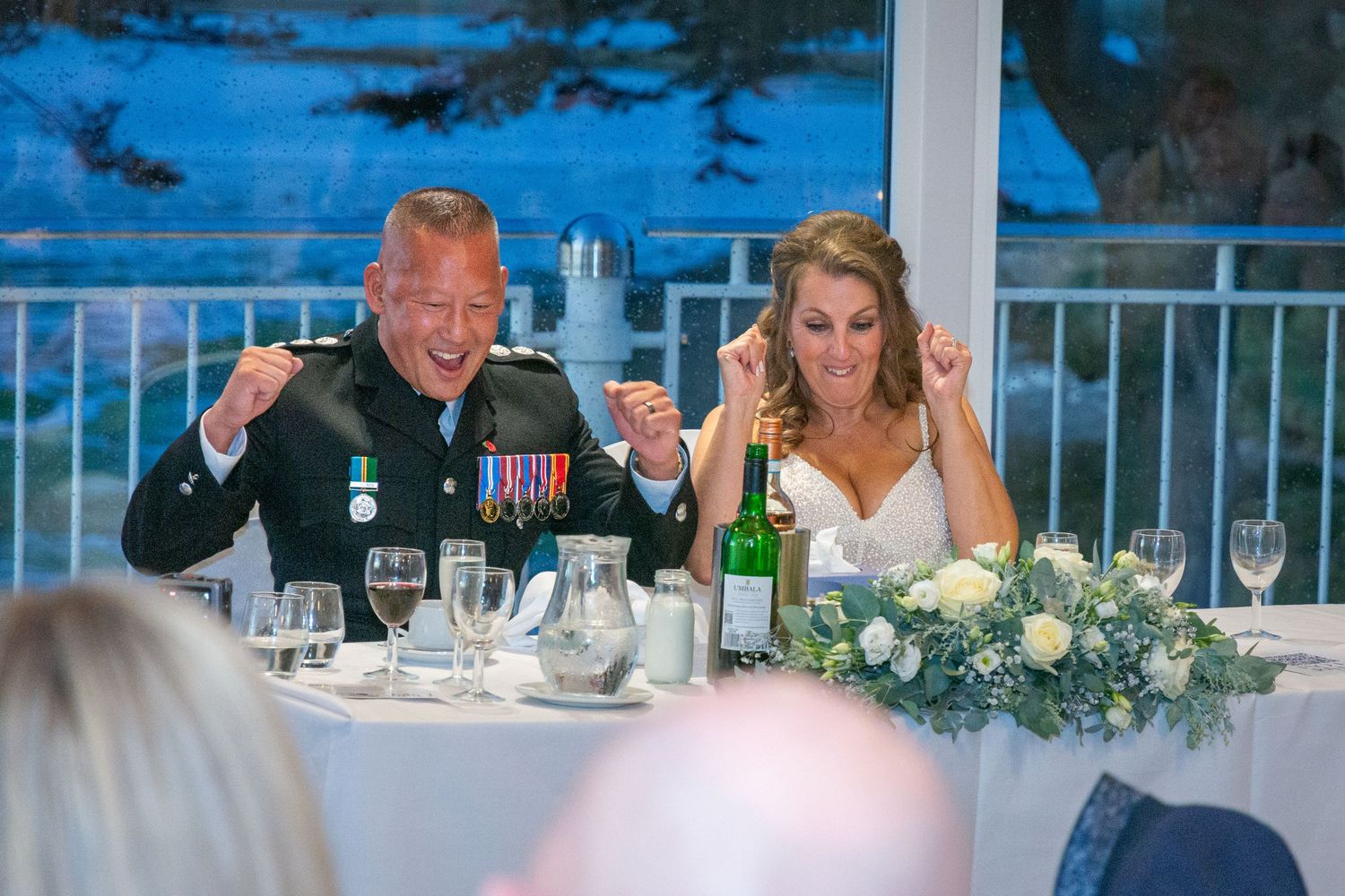 Military groom and bride laugh joyfully at wedding reception head table decorated with white roses.