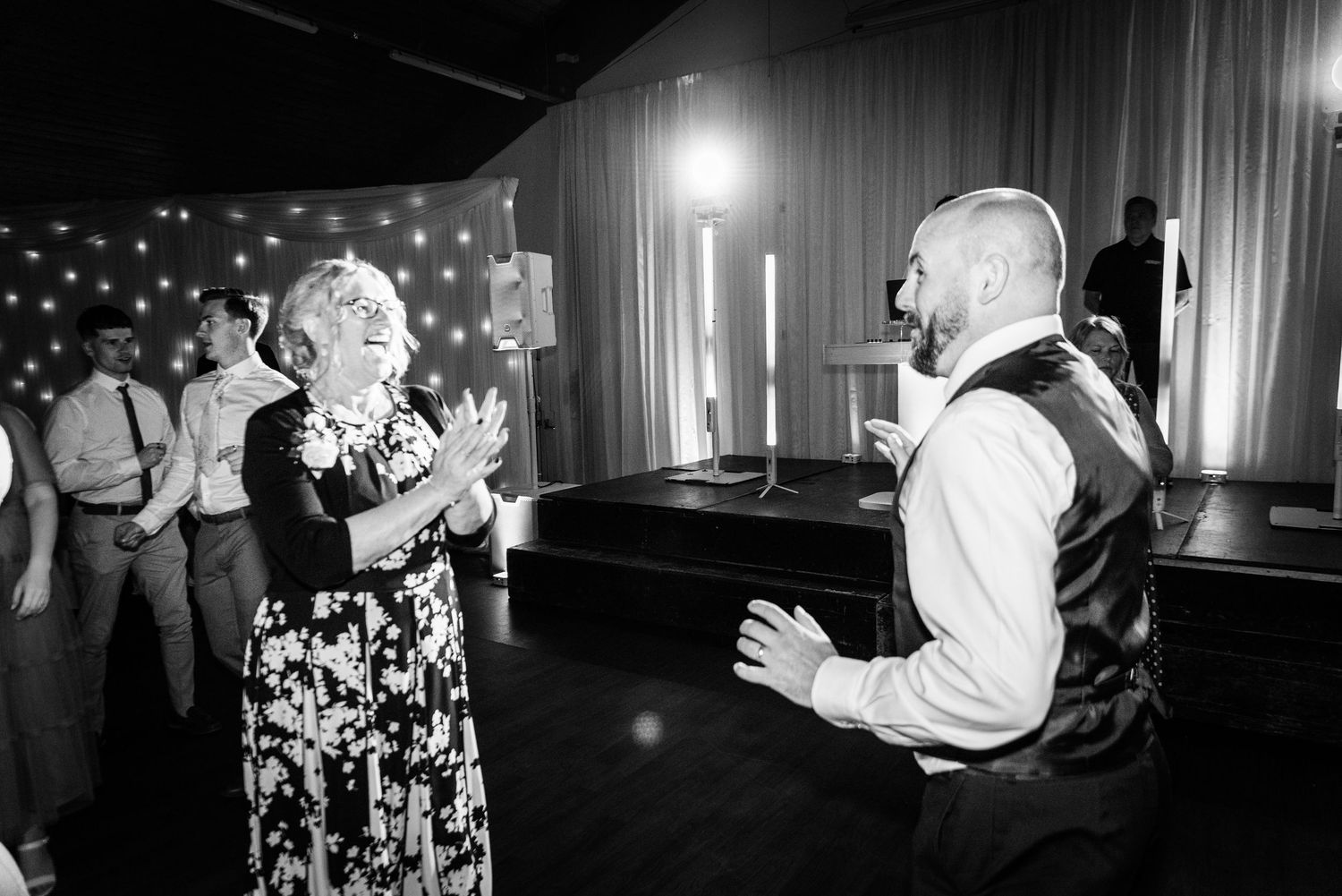 Black and white photo of groom dancing with older woman at wedding reception with bokeh lights.