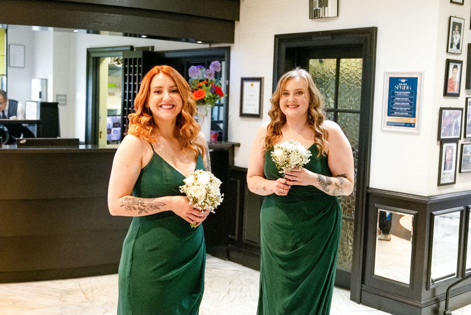Two bridesmaids in emerald green gowns holding white bouquets smile in an elegant hotel lobby.
