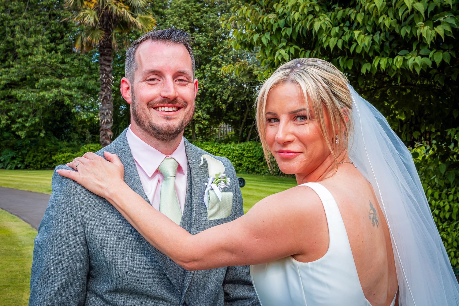 A newlywed couple poses together outdoors in wedding attire with lush green foliage in the background.