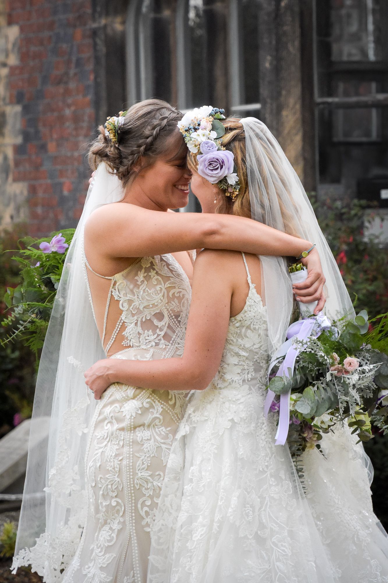 Two brides in lace gowns and floral crowns embrace tenderly outside a historic stone building.