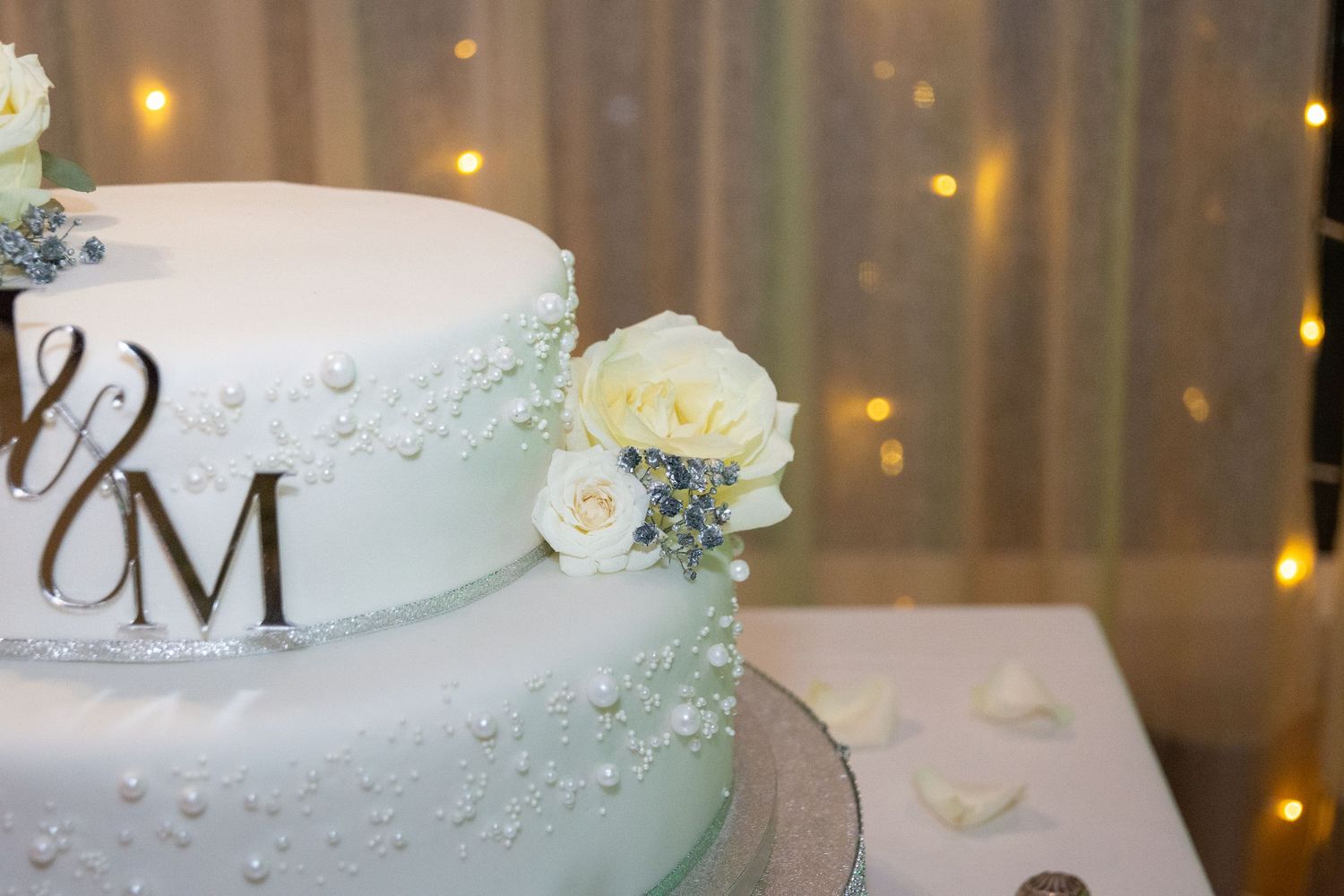 Elegant two-tier white wedding cake with pearl details, white roses, and decorative M monogram topper.