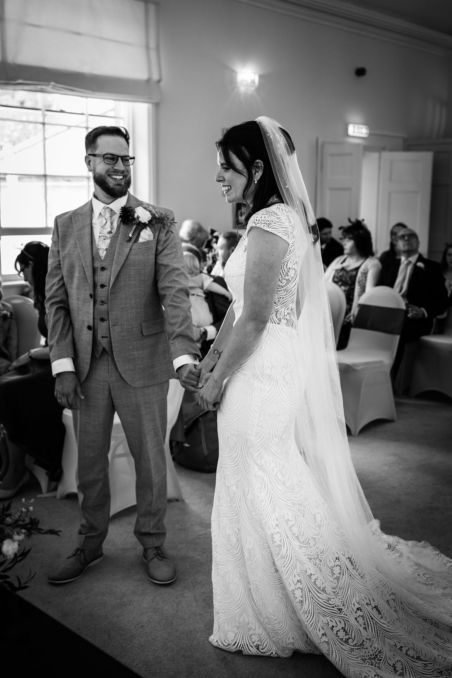 Bride and groom holding hands smiling during indoor wedding ceremony, groom in grey suit, bride in lace dress with veil.