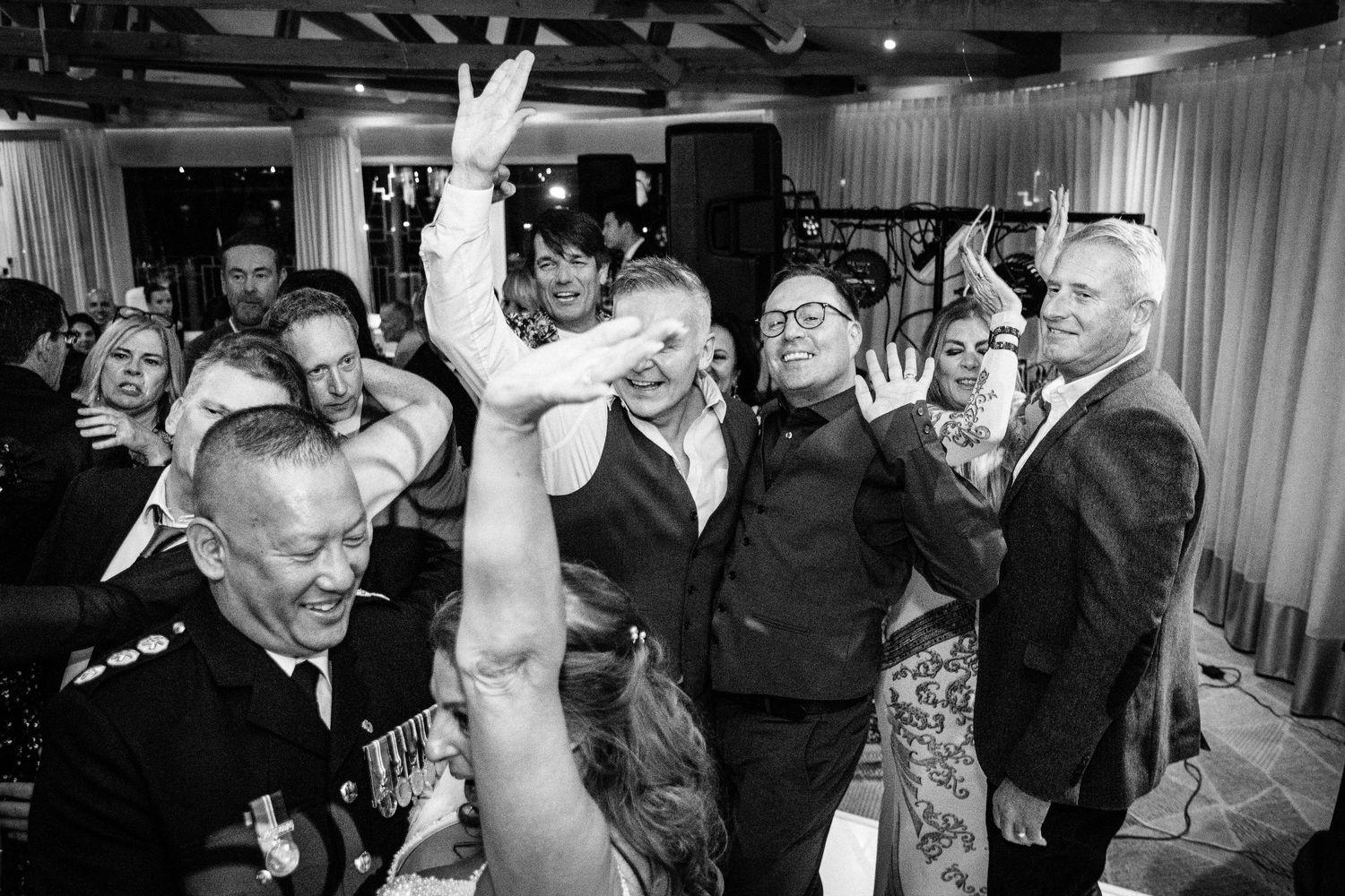 Black and white photo of joyful wedding guests dancing and celebrating together at a lively indoor reception party.