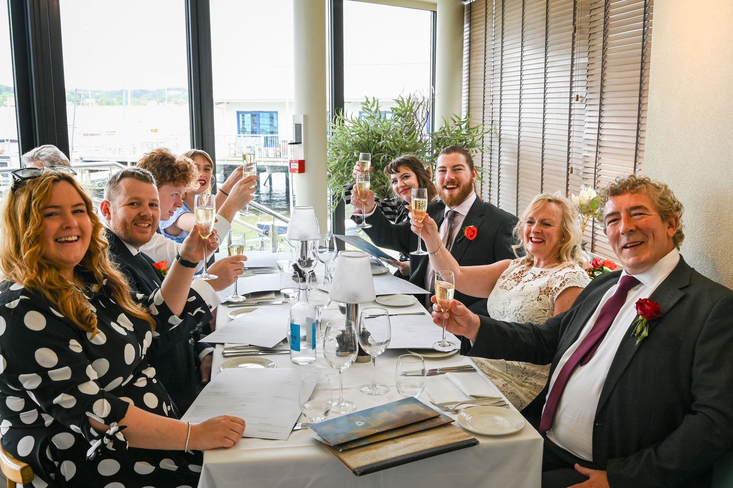 Happy wedding party guests raising glasses in a toast at a seaside restaurant reception venue.