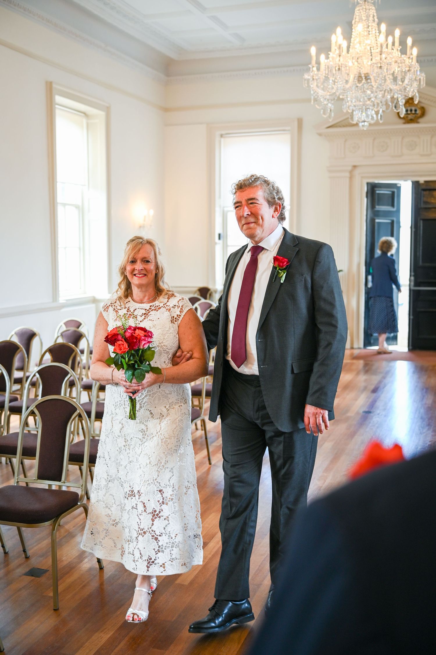Bride and groom walking down the aisle in an elegant white room with chandelier during wedding ceremony.