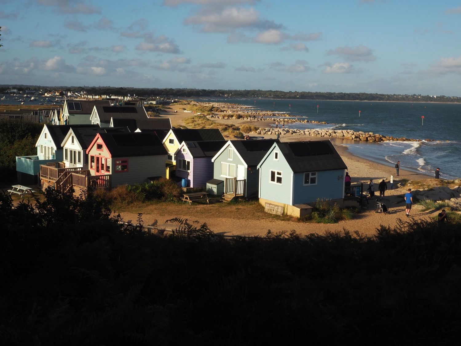 Colorful beach huts on a sunny hillside overlooking a sandy shoreline and calm blue sea.