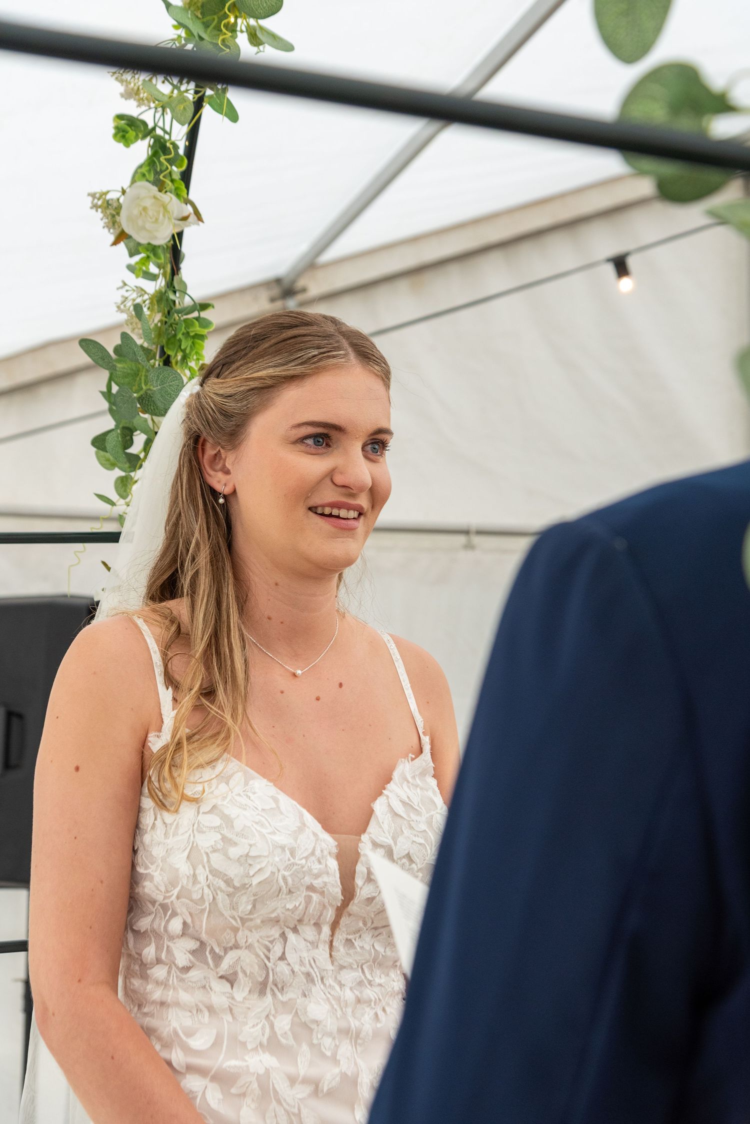 Smiling bride in a white floral wedding dress and veil standing at the altar under a floral arch.
