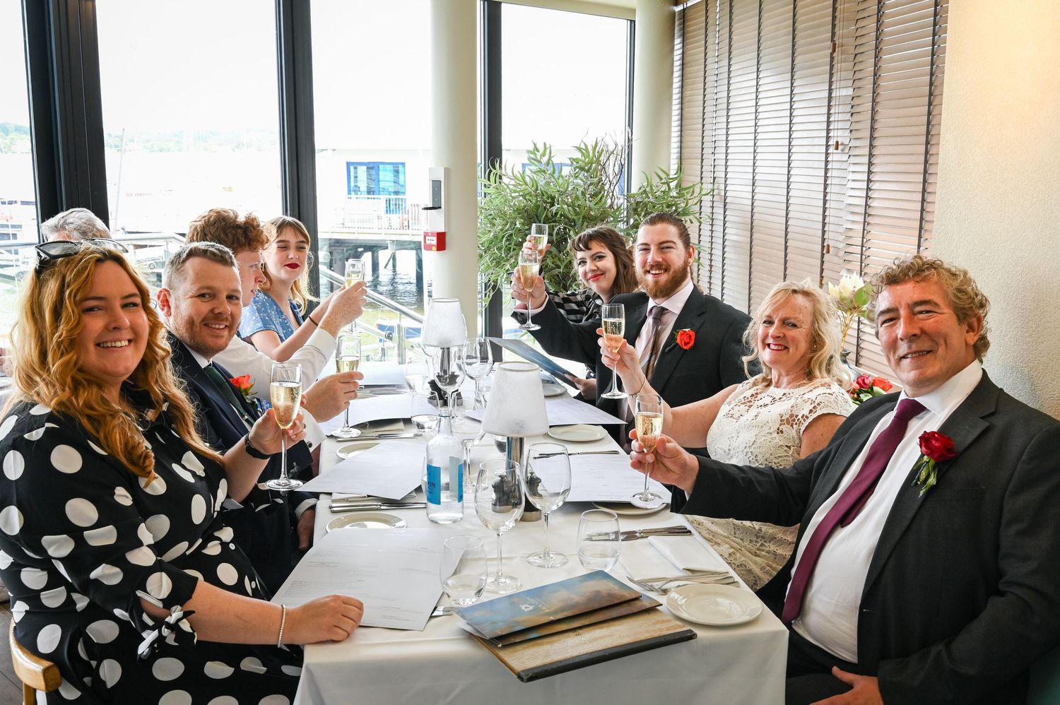 Wedding party guests raising champagne glasses in a toast at a decorated dining table near large windows.