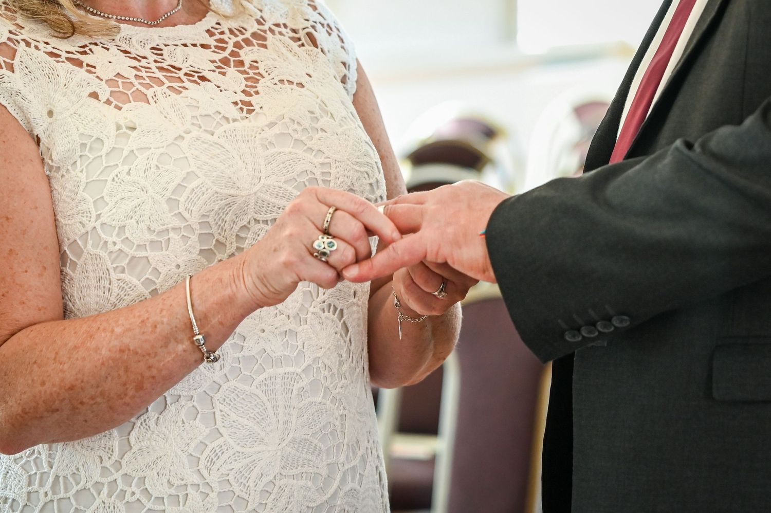 Bride in white lace dress placing wedding ring on groom's finger during ceremony.