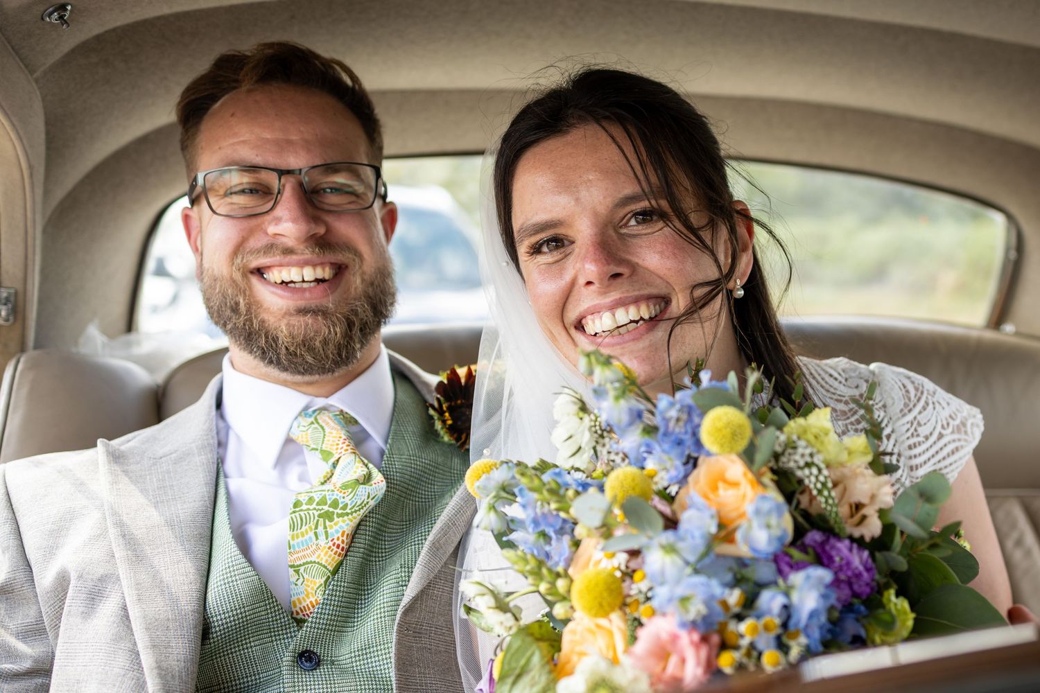 A cheerful couple sits in a car with fresh spring flowers including yellow daffodils and blue delphinium.