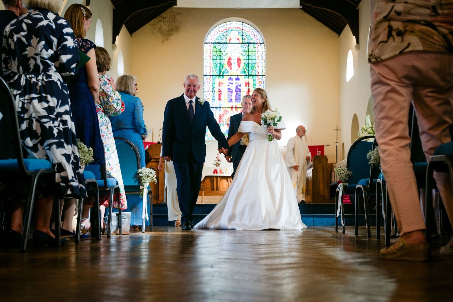 Bride walks down church aisle with escort past seated guests toward stained glass window altar.