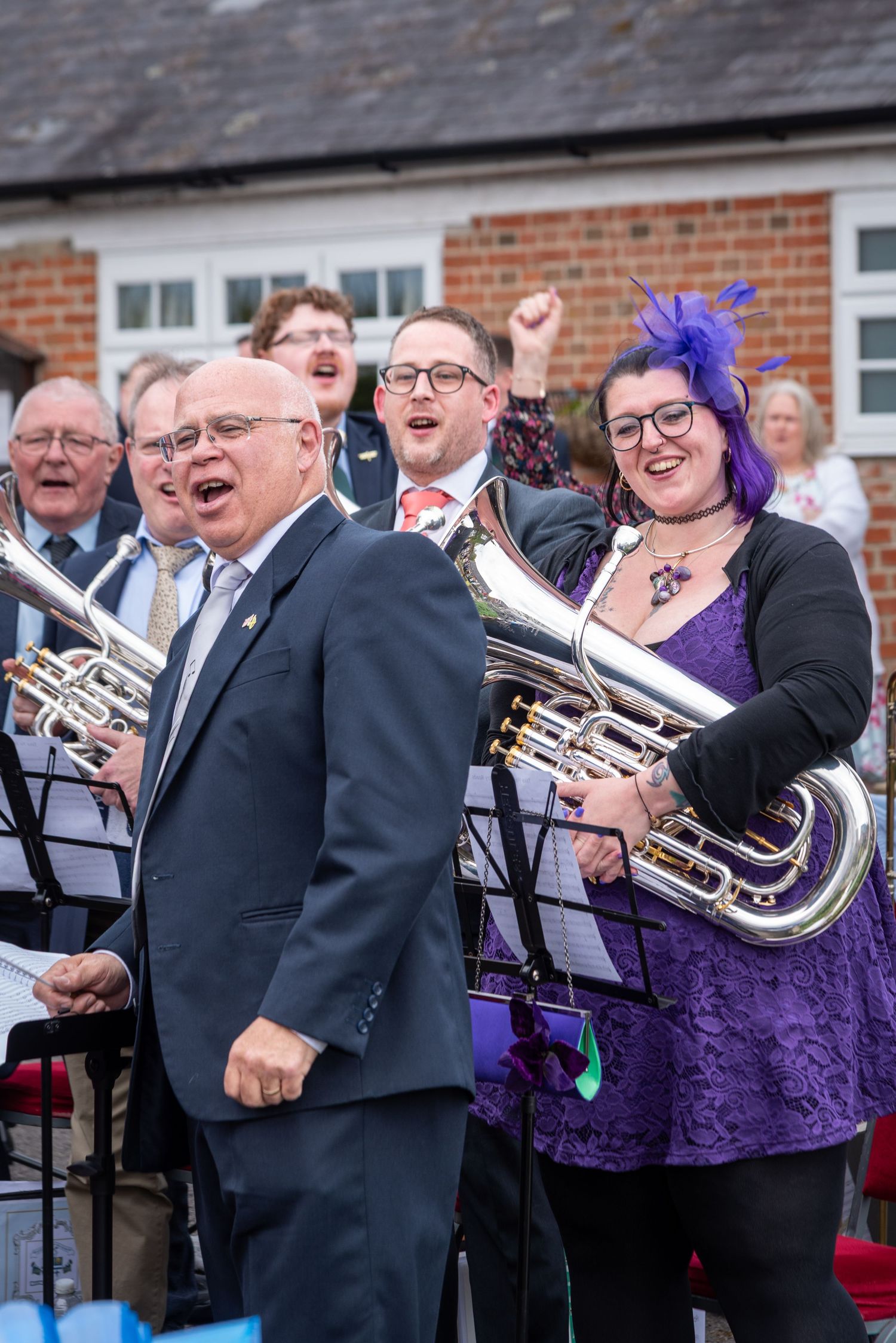 Brass band musicians in purple outfits perform outdoors at a community event, smiling and singing joyfully.
