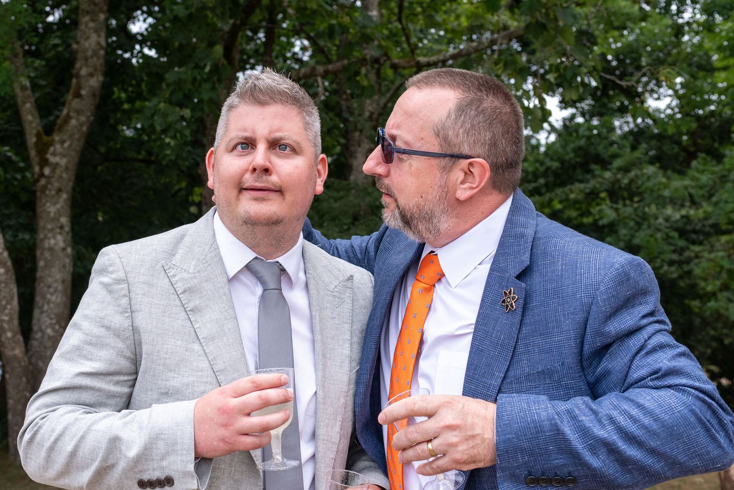 Two men in suits holding drinks outdoors at a formal event, surrounded by lush green trees.