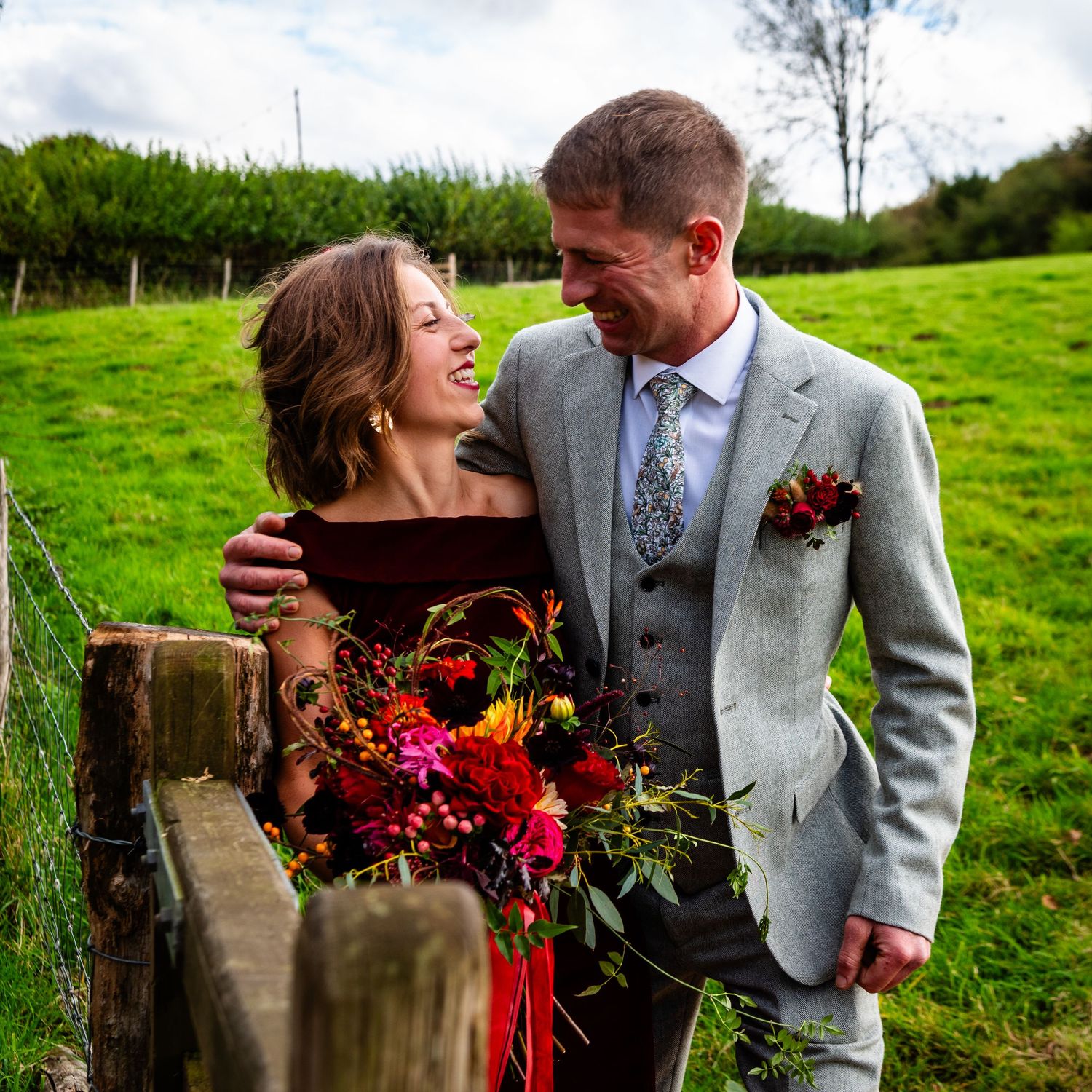 A smiling bride in burgundy dress holds colorful bouquet beside groom in gray suit at a rustic outdoor wedding.