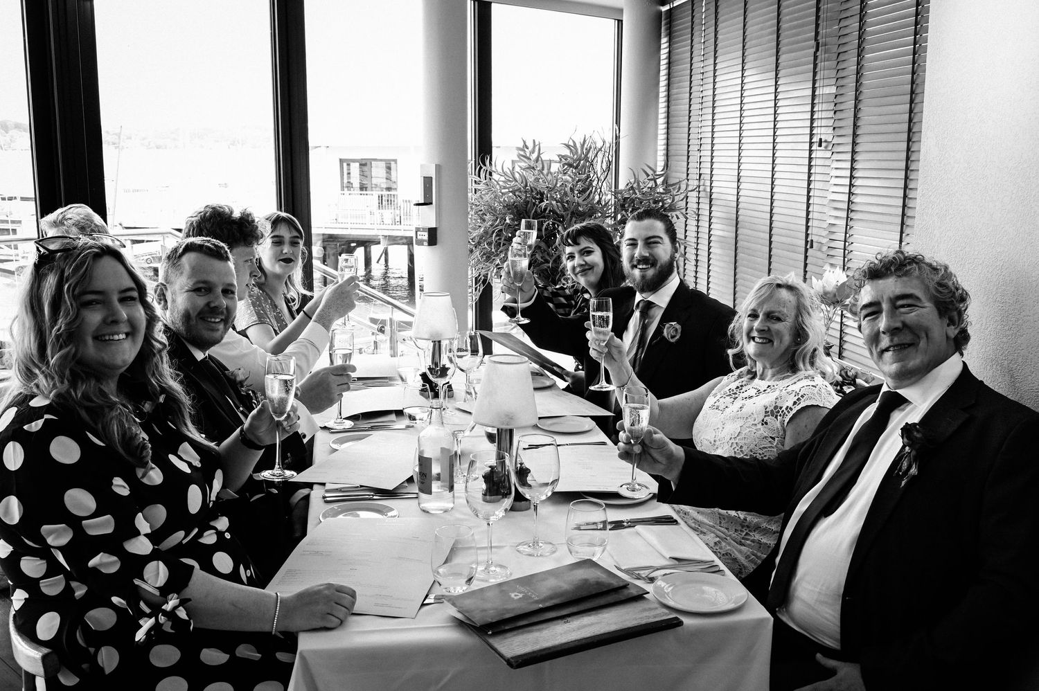 Group of guests raising champagne glasses in a toast at a wedding reception dinner table, black and white photo.