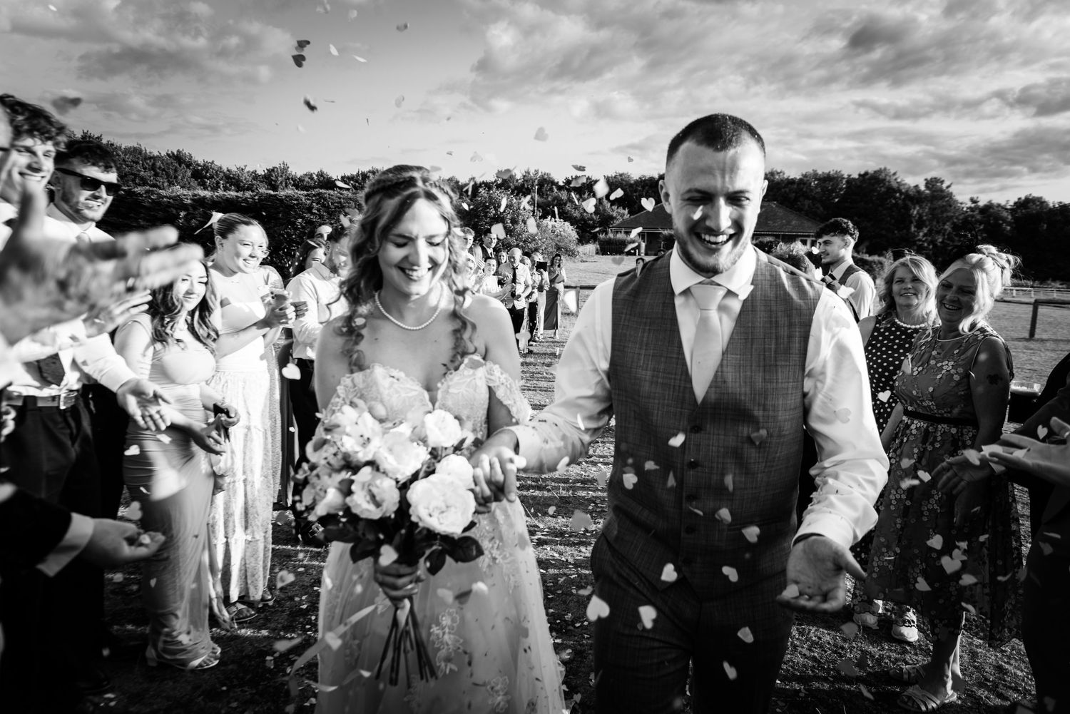 Black and white photo of a joyful bride and groom walking through confetti shower at outdoor wedding ceremony.