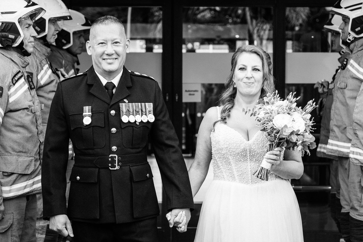 A smiling couple in military uniform and wedding dress exit a ceremony through a guard of honor of firefighters.