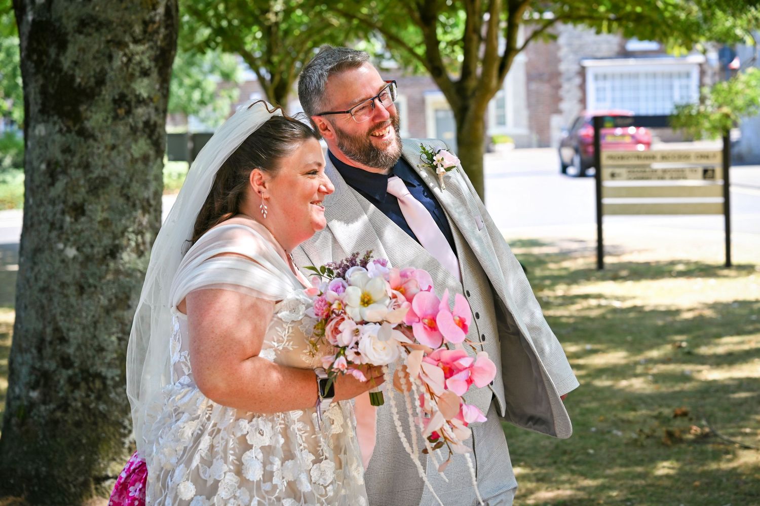 Bride in white gown and veil holds pink bouquet beside groom in gray suit, smiling outdoors under trees.