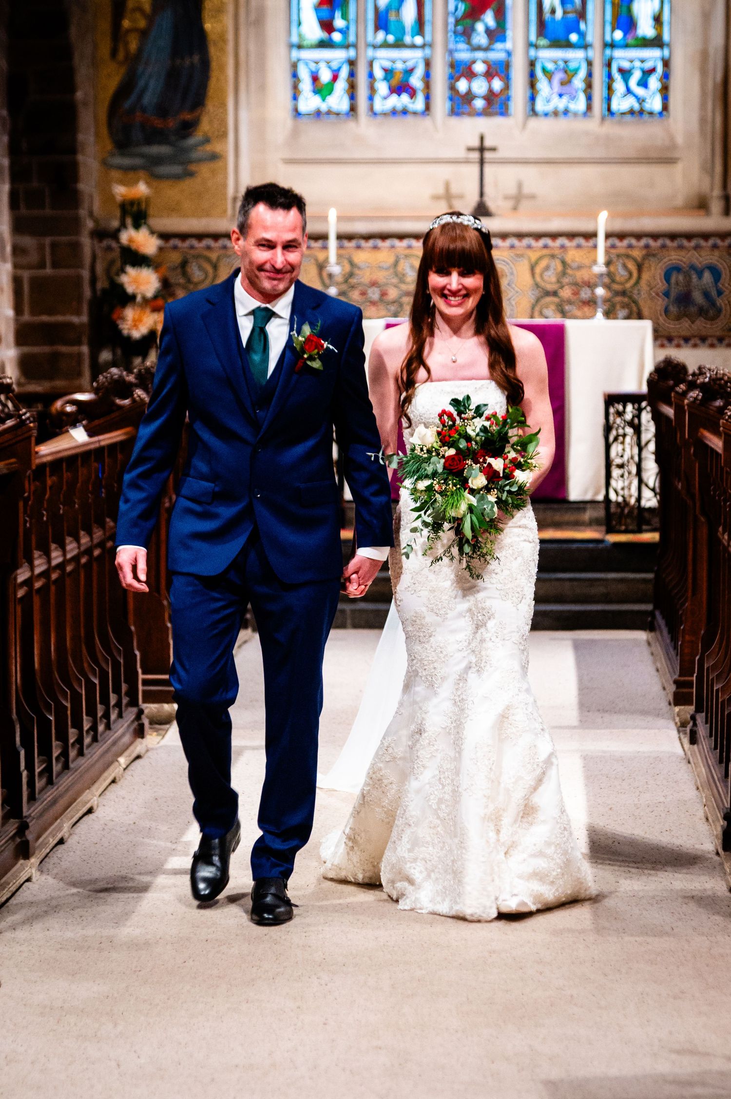 Bride and groom walk down church aisle after wedding ceremony, bride holding lush bouquet, groom in navy suit.