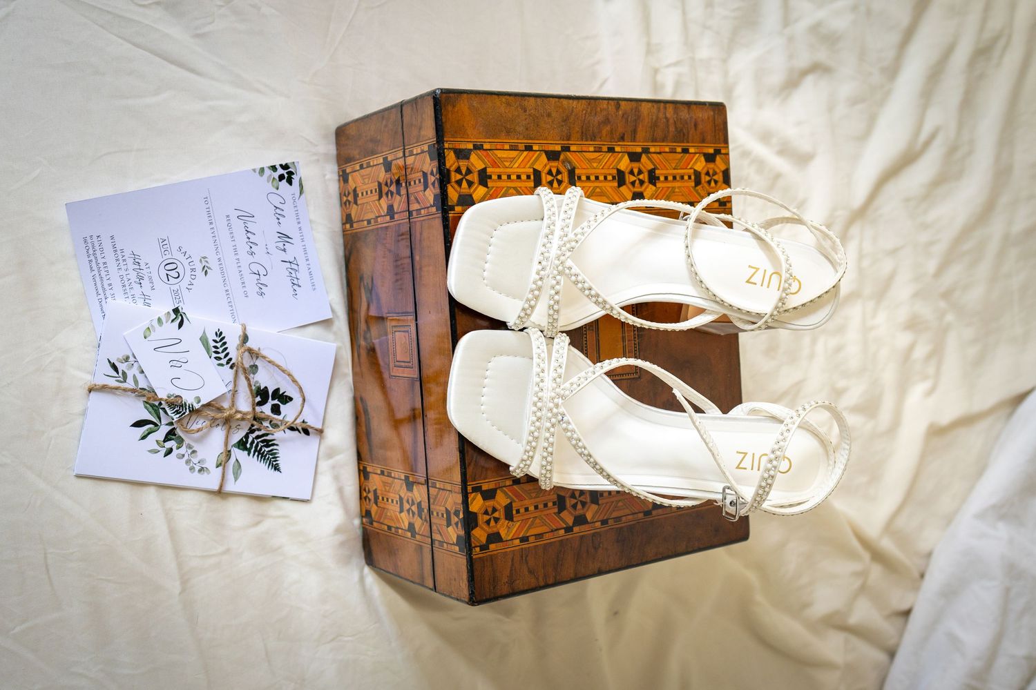 White pearl strappy bridal sandals displayed on a wooden box beside wedding invitation on white fabric.