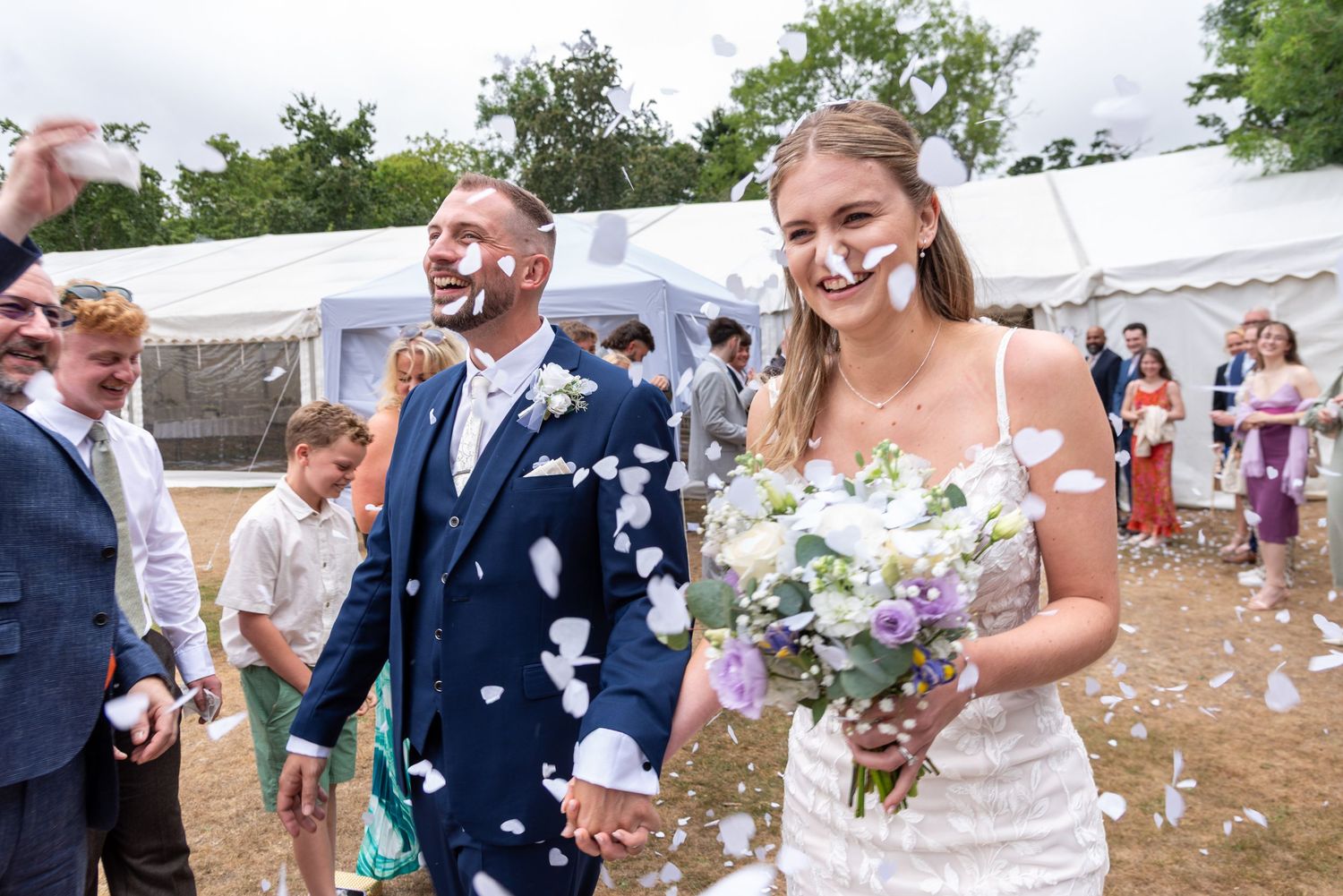 Bride and groom walk hand-in-hand through confetti shower at outdoor wedding celebration.