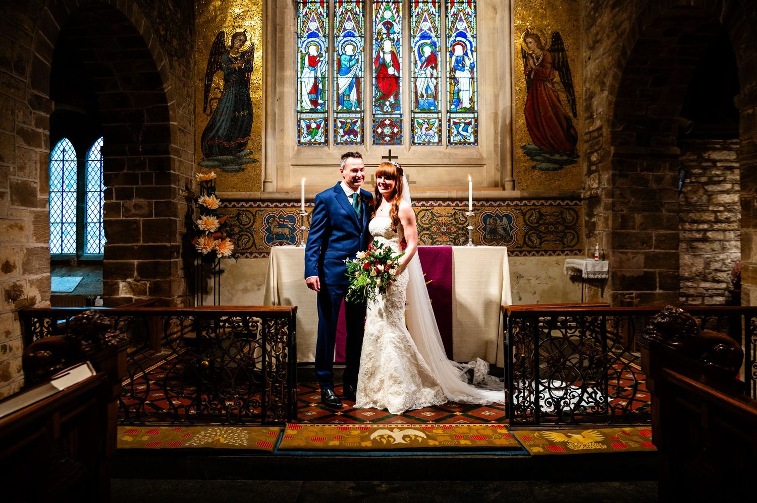 A couple stands together in a historic church with beautiful stained glass windows and stone archways in the background.