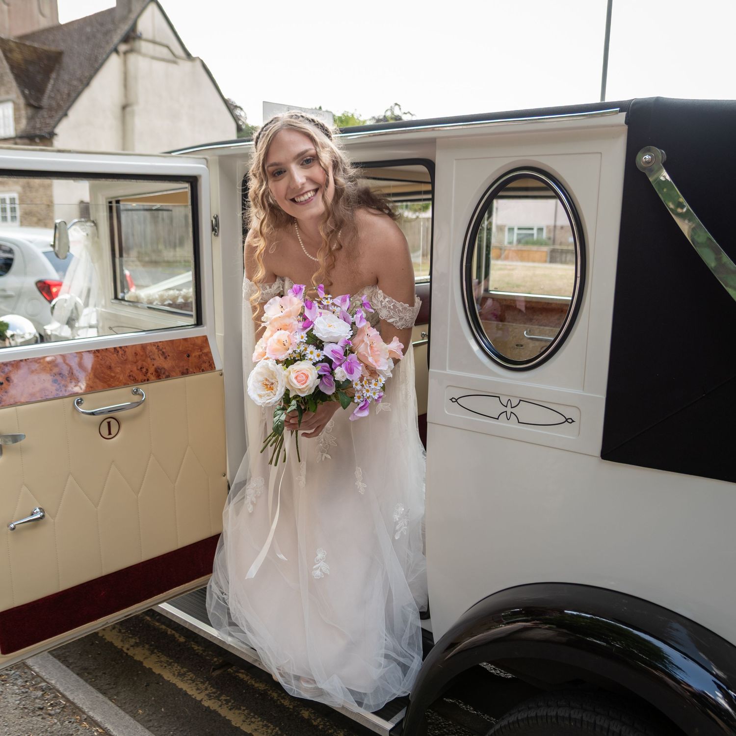Smiling bride in white off-shoulder gown holding pastel bouquet stepping out of vintage cream wedding car.