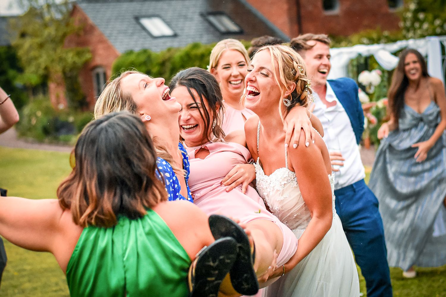 Joyful wedding guests laugh and embrace the bride in a white dress during an outdoor celebration.