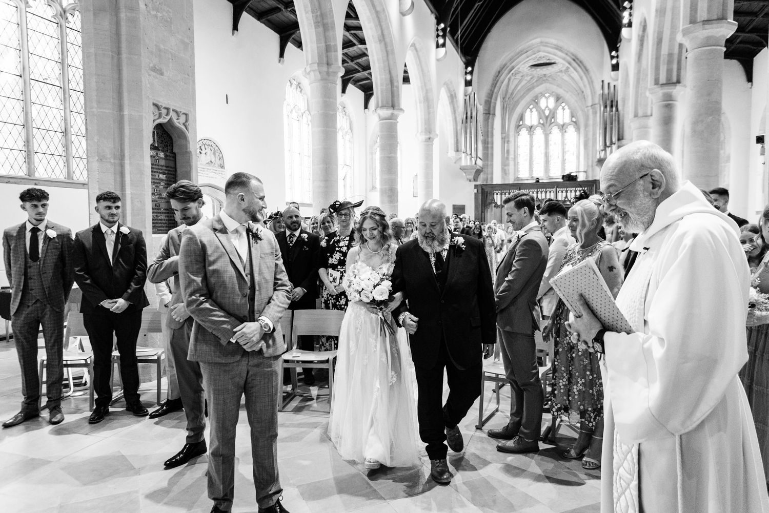 Bride walks down the aisle with her father in a historic church, as groom and officiant await at the altar.