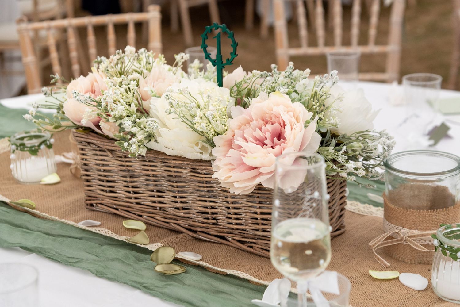 Rustic wicker basket centerpiece with pink peonies and white flowers on a burlap table runner at a wedding reception.