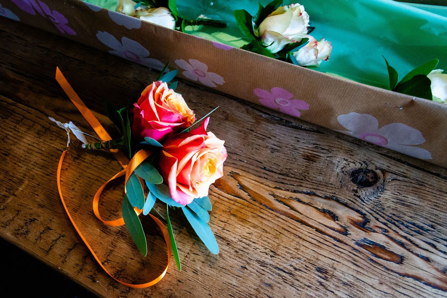 Two pink roses with orange and green ribbon lay on a rustic wooden surface beside a turquoise fabric in sunlight.