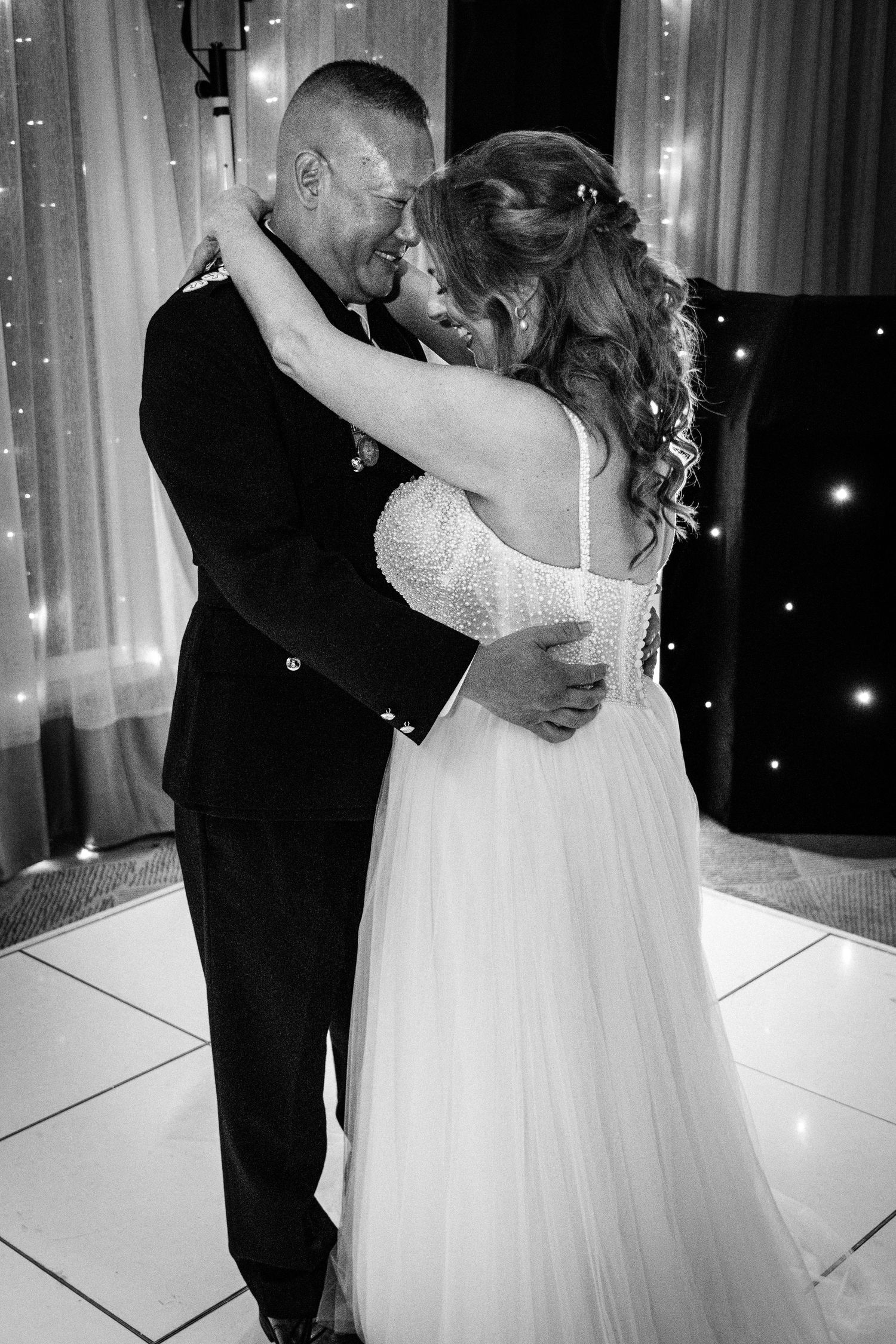 Bride and groom share romantic first dance embracing on white dancefloor with fairy lights backdrop.
