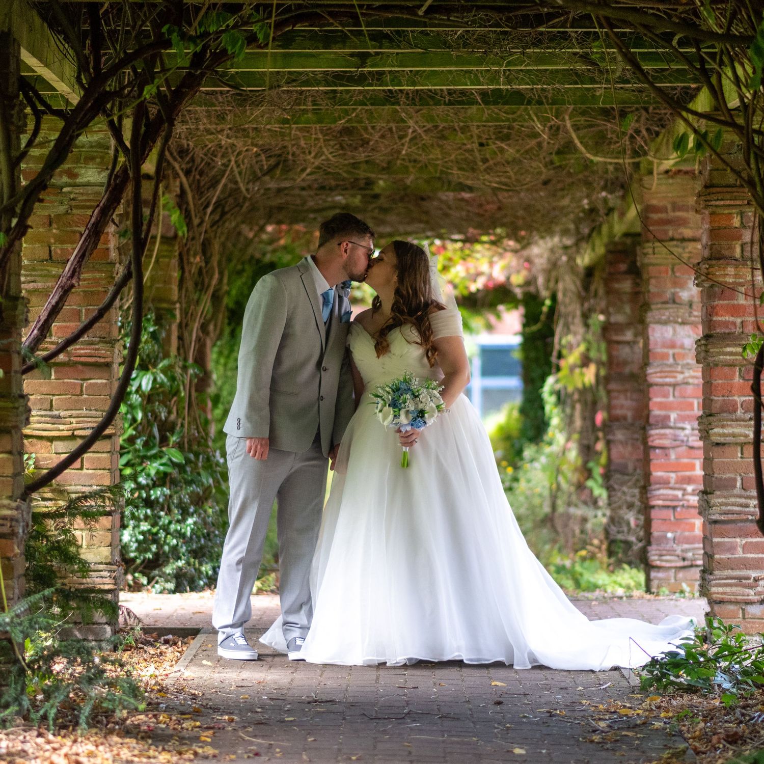 A bride and groom share a romantic kiss under a vine-covered brick archway in a lush garden setting.