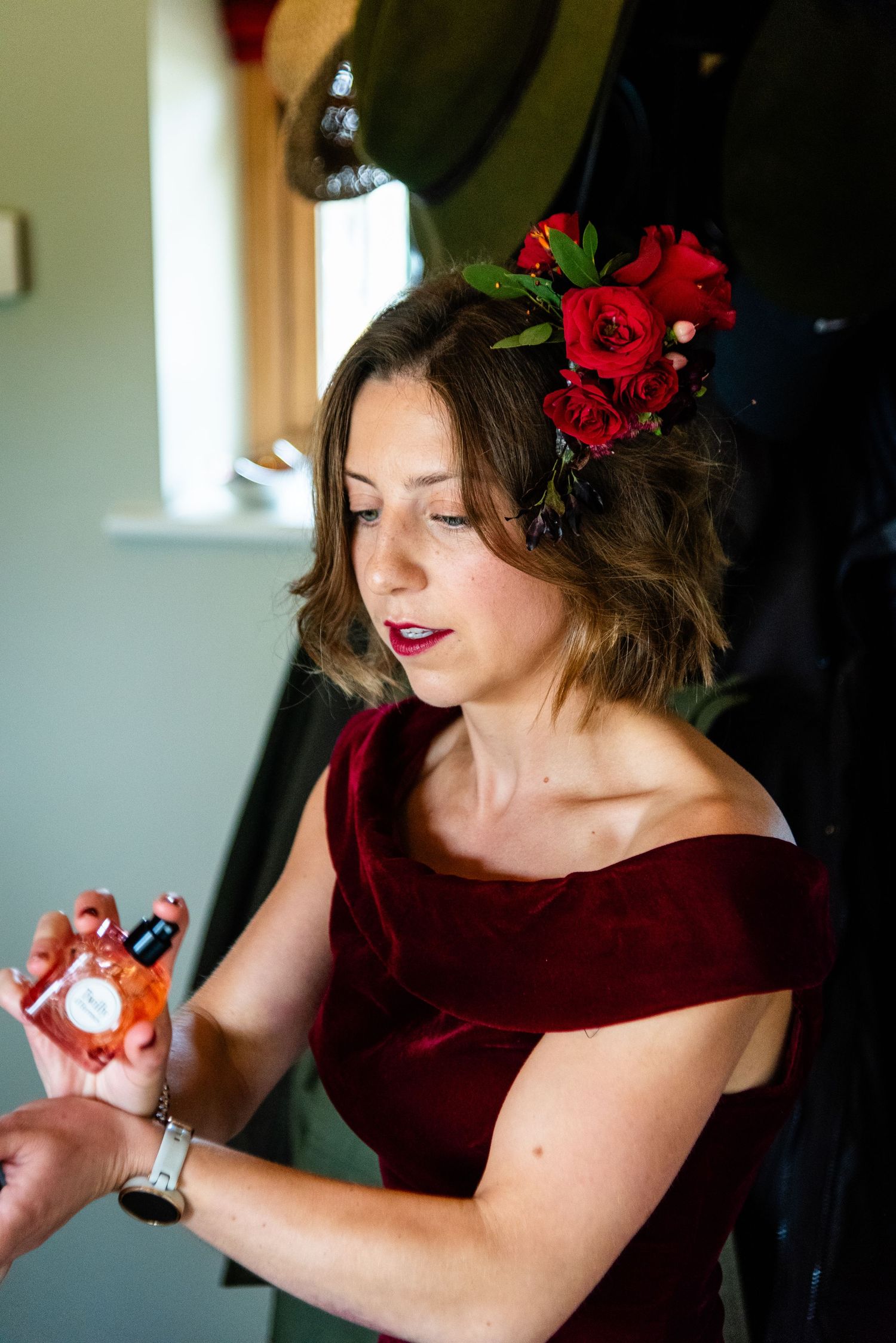 Woman in red off-shoulder dress with red roses in hair applies perfume to her wrist.