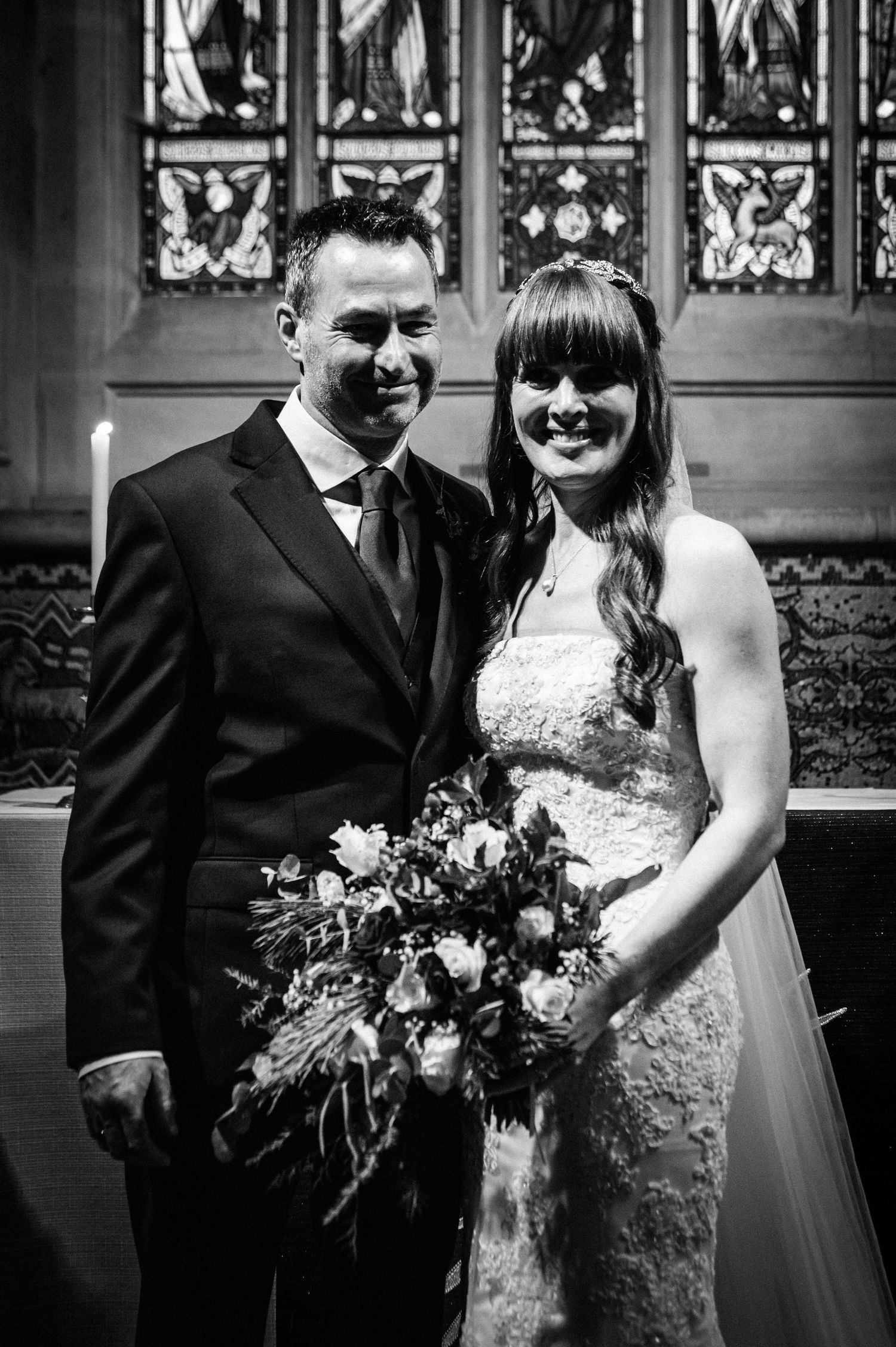 Black and white photo of a smiling bride and groom posing together in a church with stained glass windows behind them.