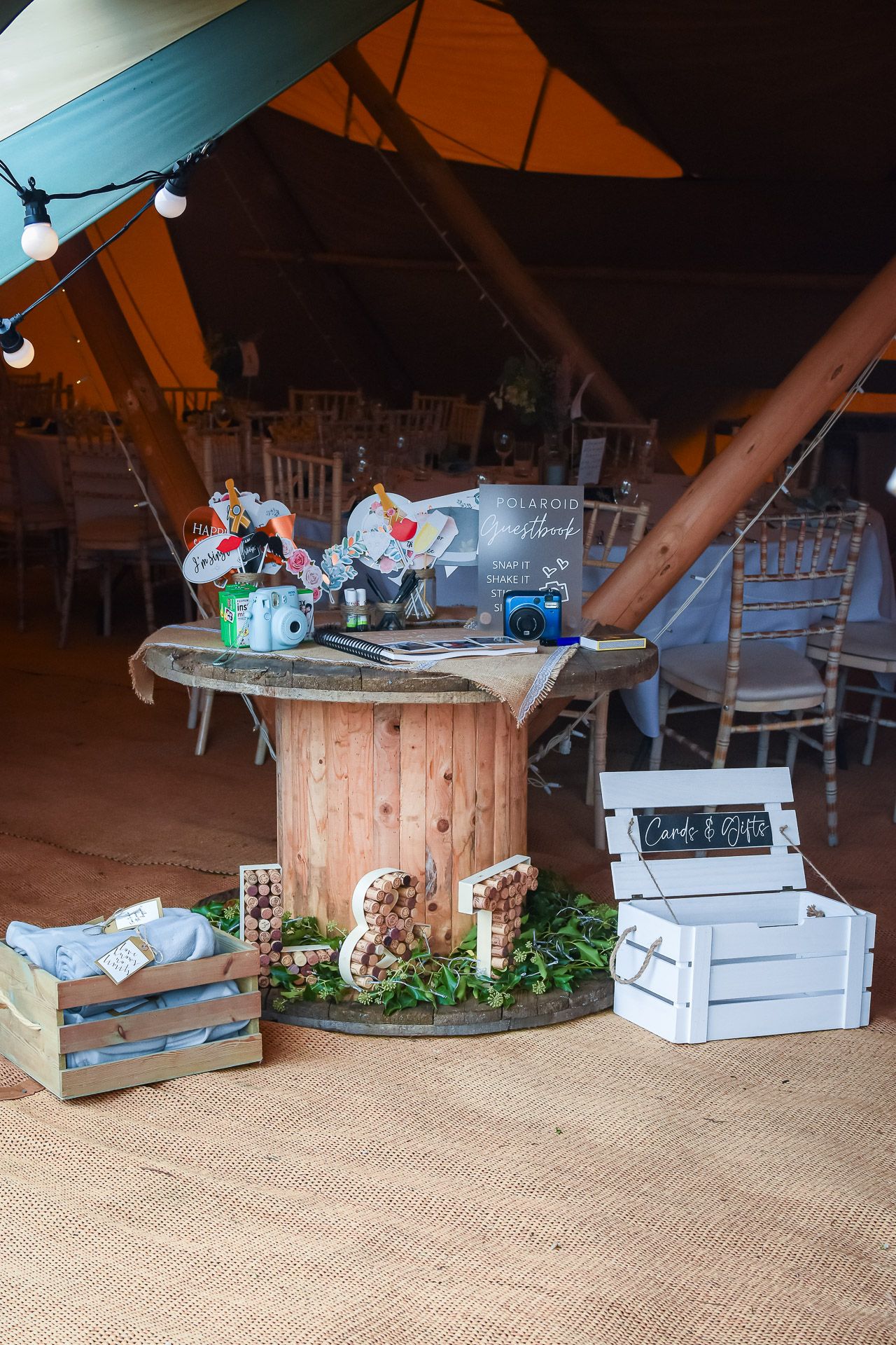 Rustic wooden spool table with decorative boxes and greenery inside a tent at an outdoor event setup.