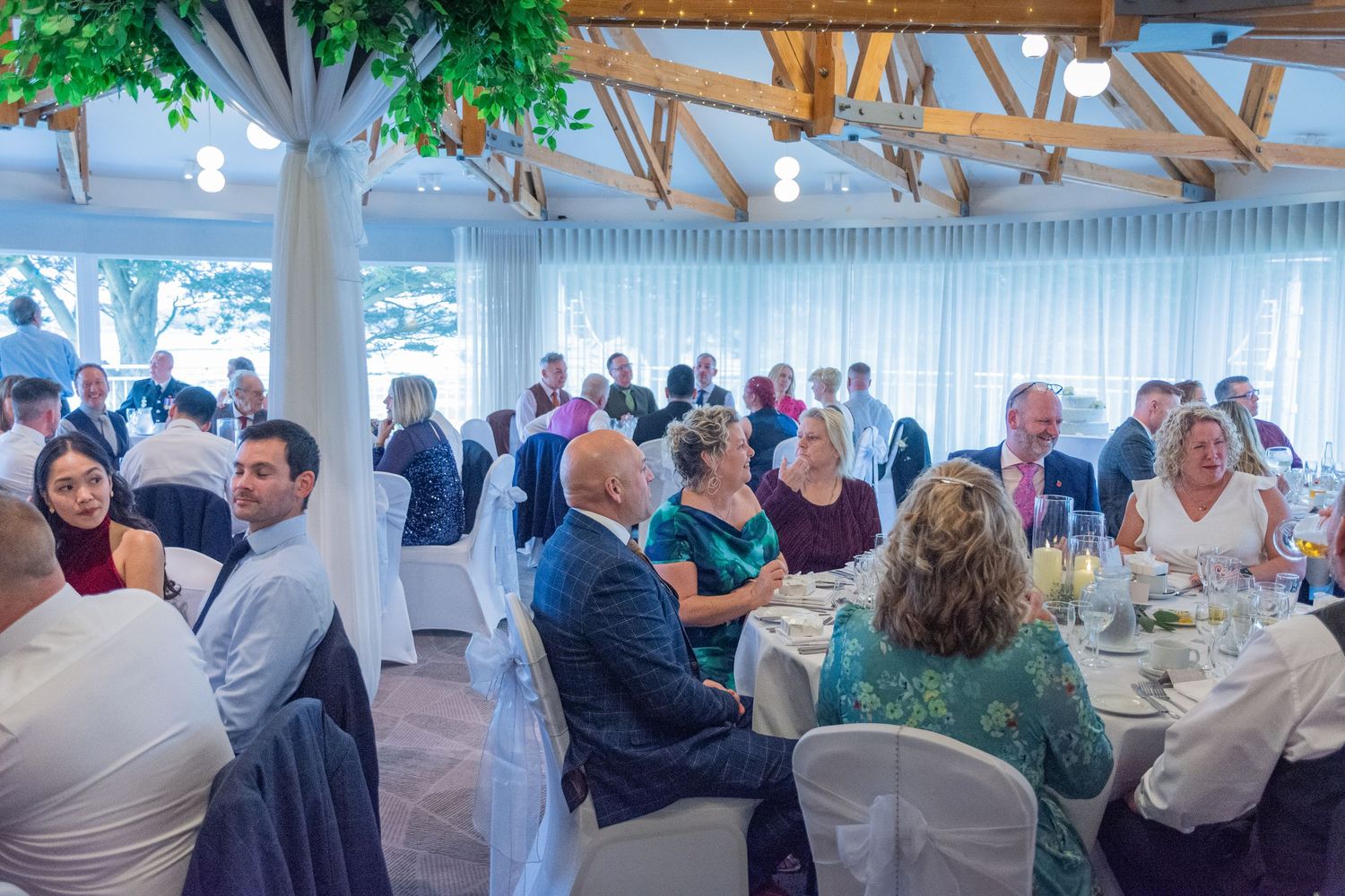 Elegant wedding reception with guests seated at round tables in a bright venue with white drapes and decorative trees.